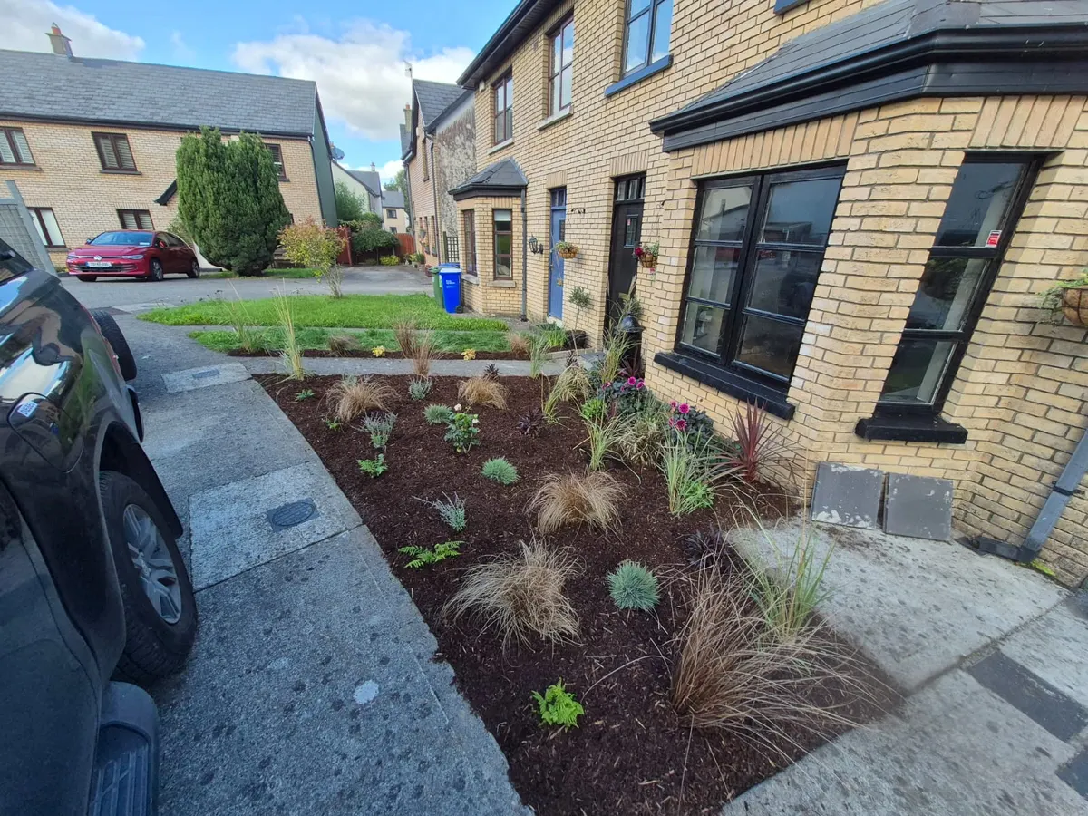Front garden bed of ornamental grasses and young shrubs in dark bark mulch along a yellow-brick terrace