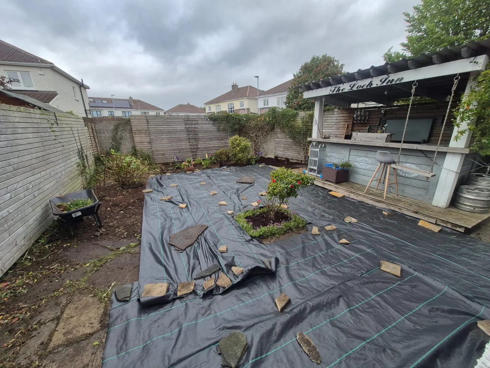 Back garden cleared and covered in weed membrane around a rose bed, next to a timber outdoor bar
