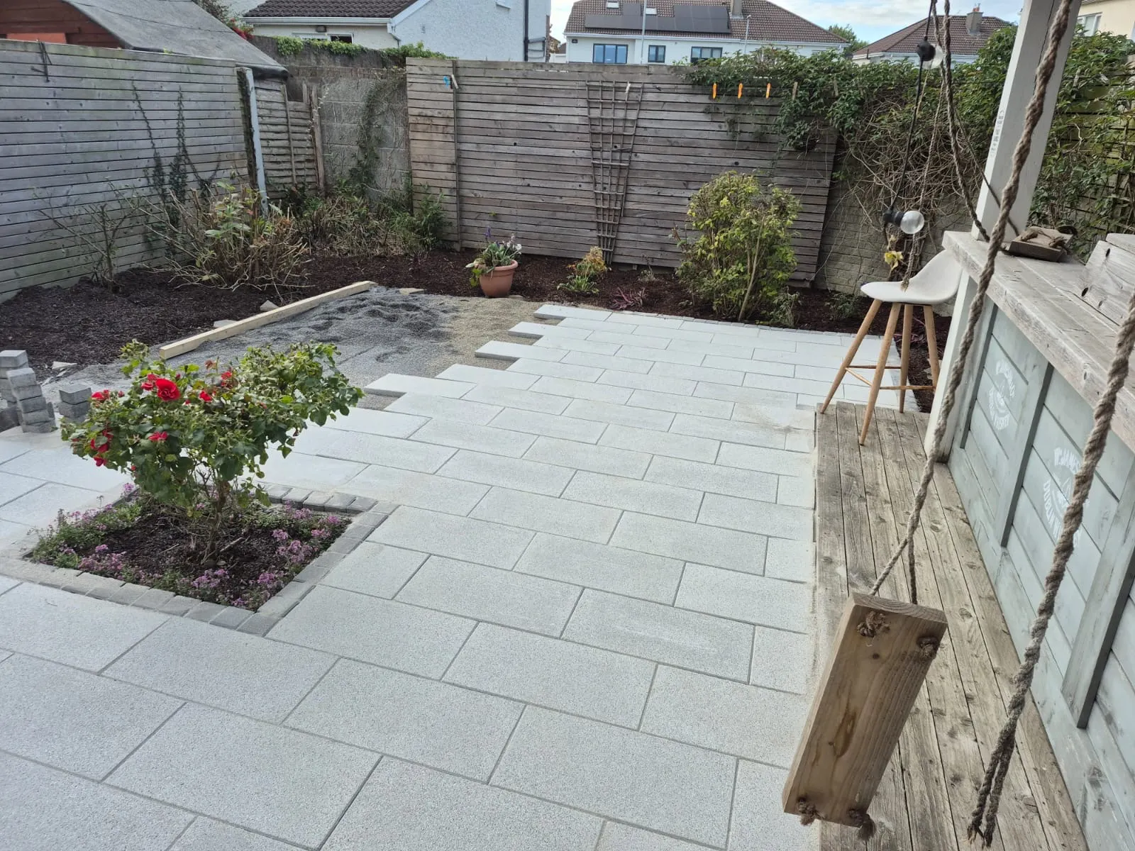 Finished pale grey porcelain patio in a back garden with a rose bed, timber bar and fenced borders