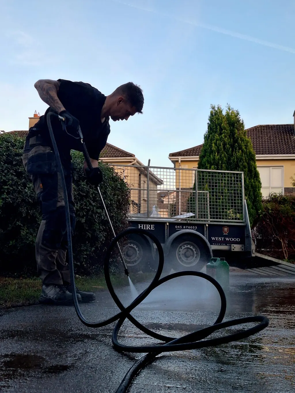 Worker pressure washing a driveway beside a hedge and trailer