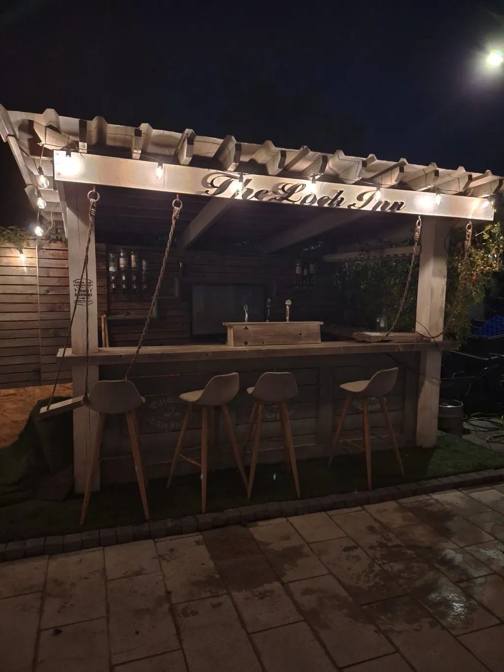 Timber outdoor bar and pergola with warm lights in a paved garden at night